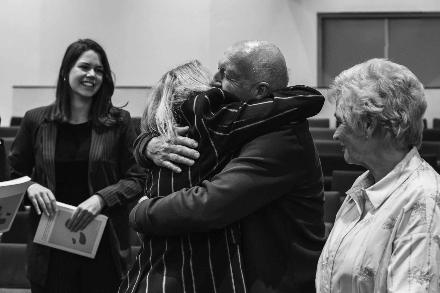Black and white photo of people hugging at a formal gathering, with smiling woman holding papers nearby.
