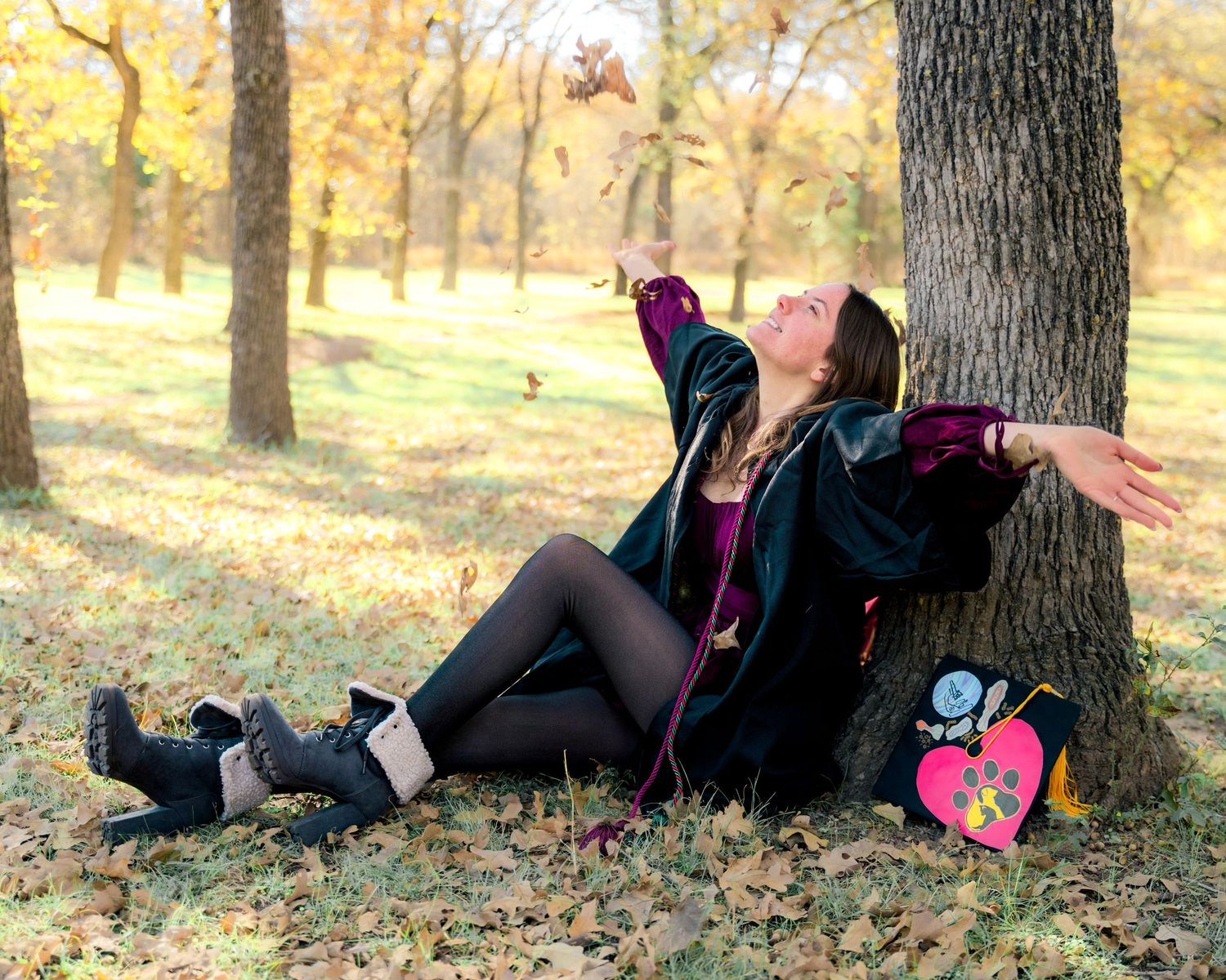 Graduate in black robe relaxing against tree trunk in autumn park with fallen leaves.