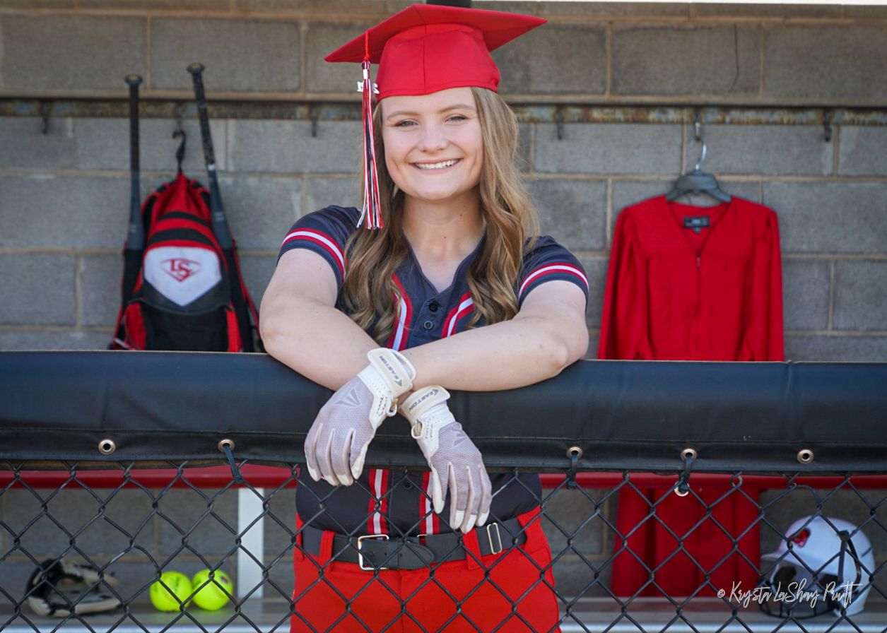 Graduate in red cap and gown posing by red fence with sports equipment visible in background.