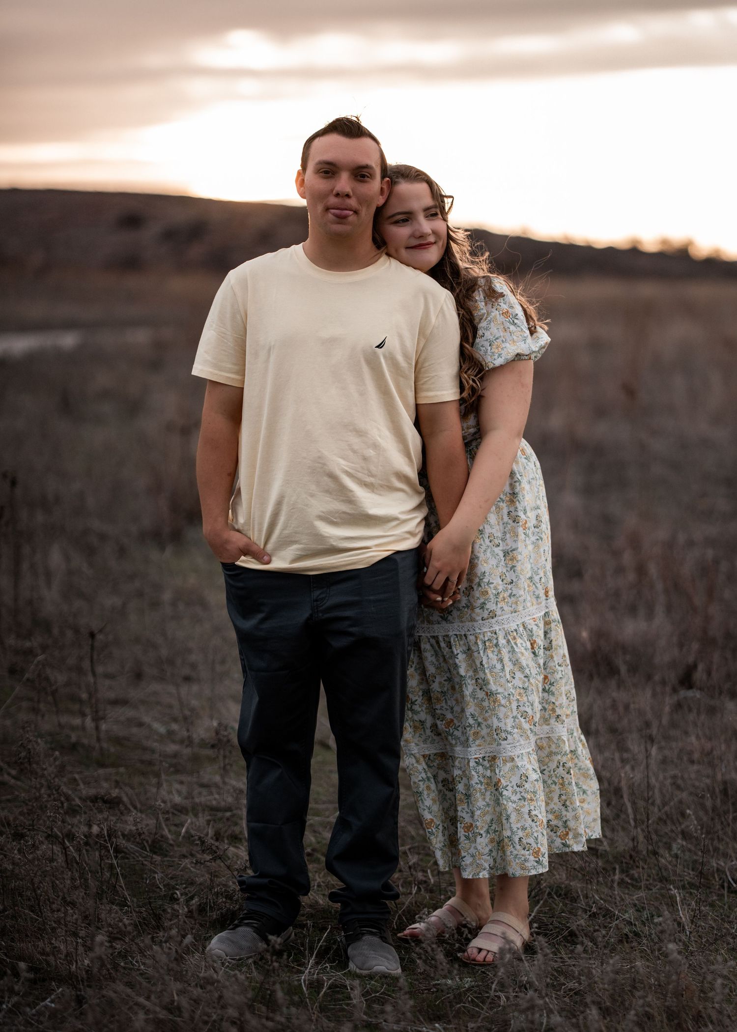 Couple poses together in casual attire during sunset in a rustic field setting.