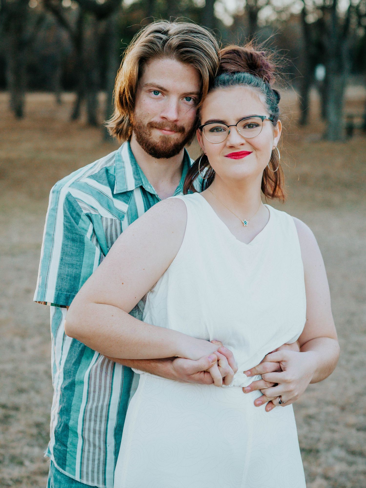 Couple poses together in casual outfits against rustic outdoor background.