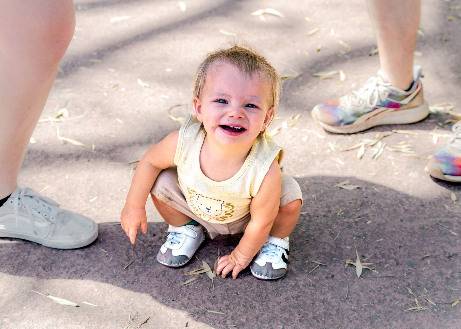 Low angle view of feet and a crawling baby on pavement in dappled sunlight.