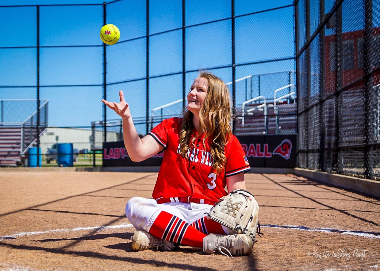 Softball player in red uniform sliding and diving for the ball on dirt field.