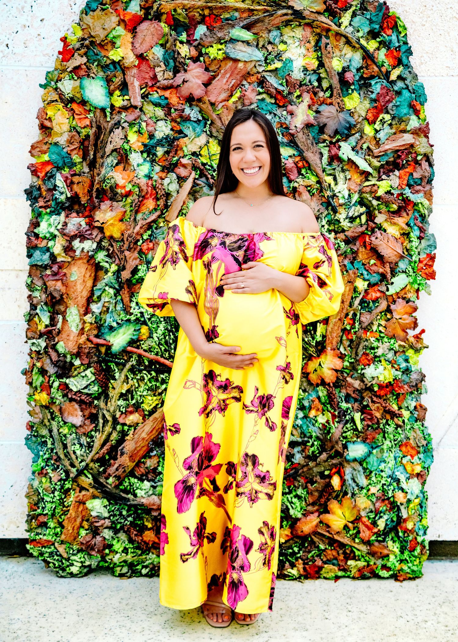 A bright yellow floral off-shoulder maternity dress is showcased against a decorative wall of colorful foliage.