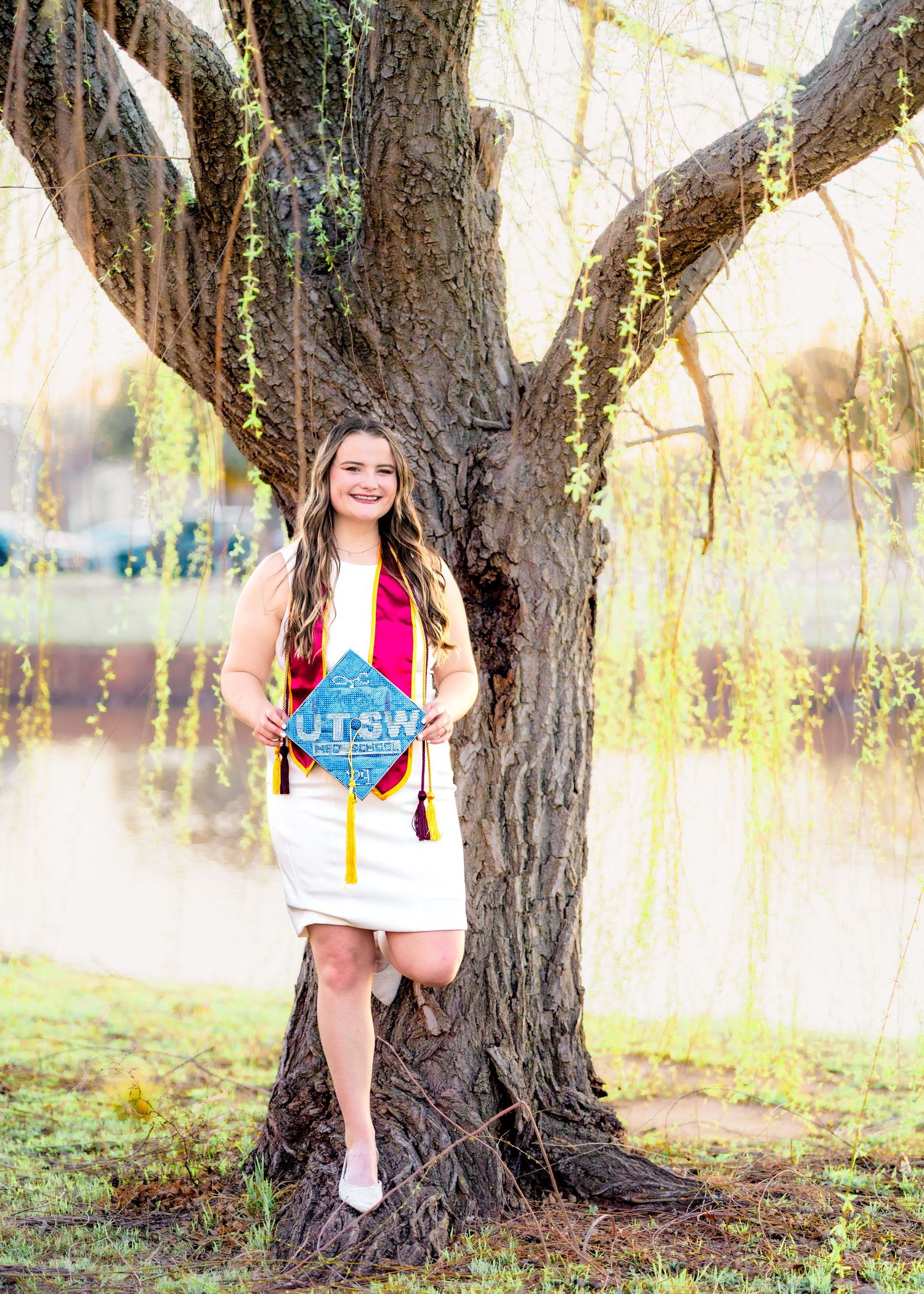 Graduate holding decorated cap poses among trees in natural outdoor setting.