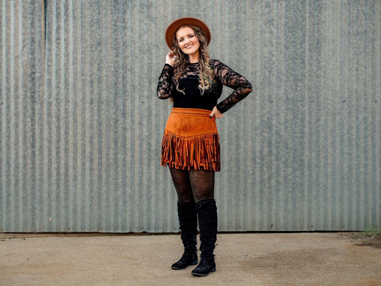 Western-inspired outfit featuring an orange fringe skirt, black lace top, and tall boots against a metal wall.