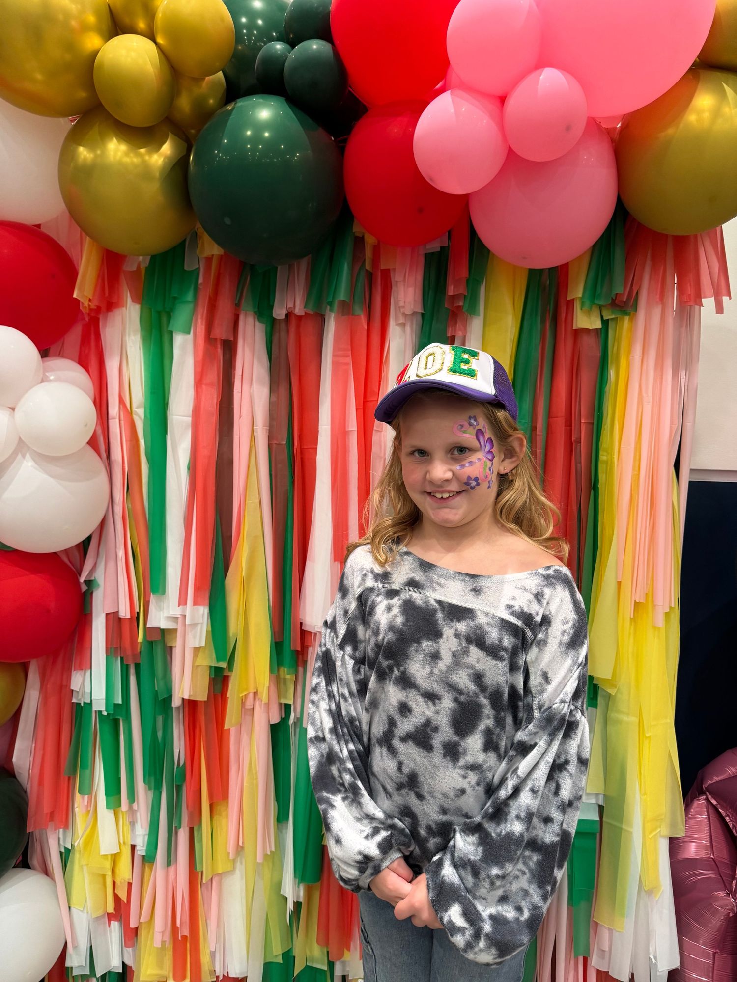 Person in tie-dye shirt and baseball cap poses in front of colorful balloon and streamer backdrop.