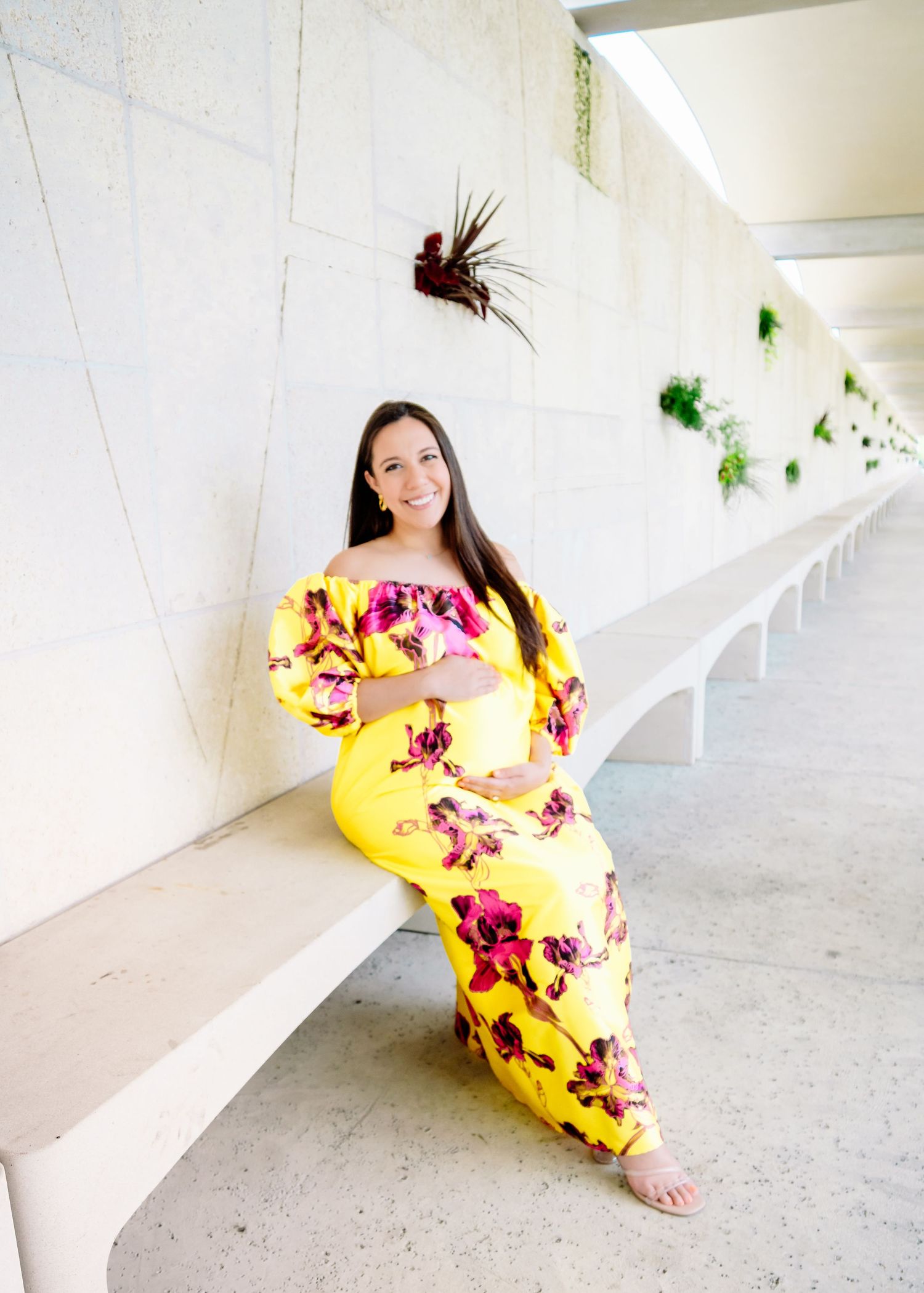 Woman in bright yellow floral dress poses gracefully in white hallway for maternity photos.