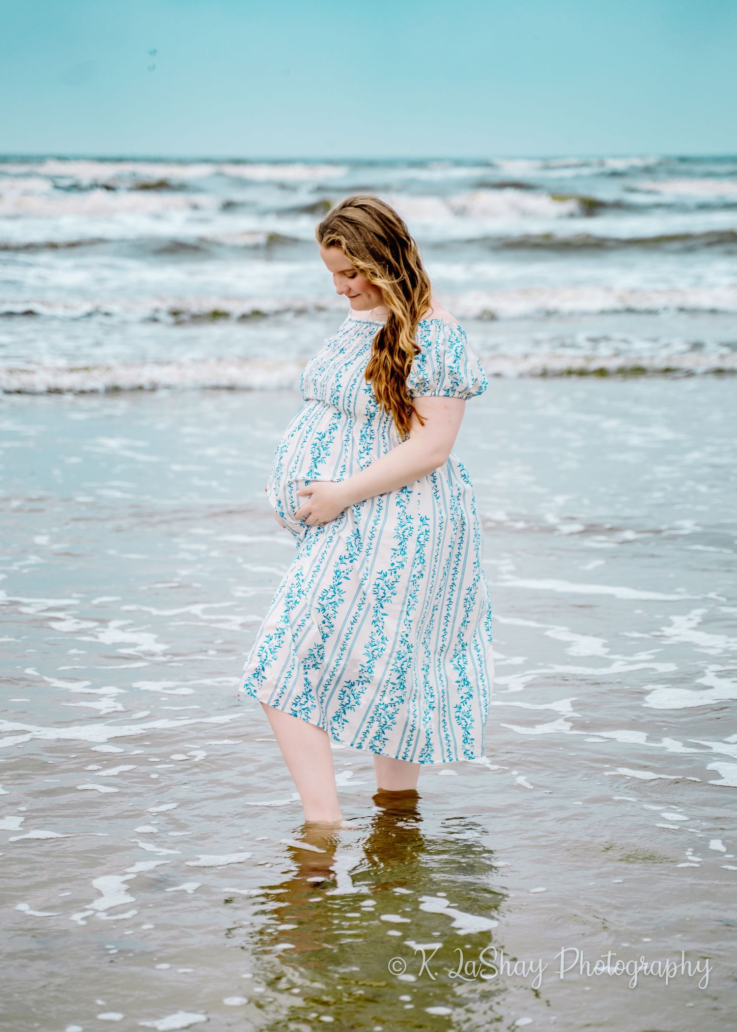 A pregnant woman in a flowing blue striped dress stands in gentle ocean waves at the beach.