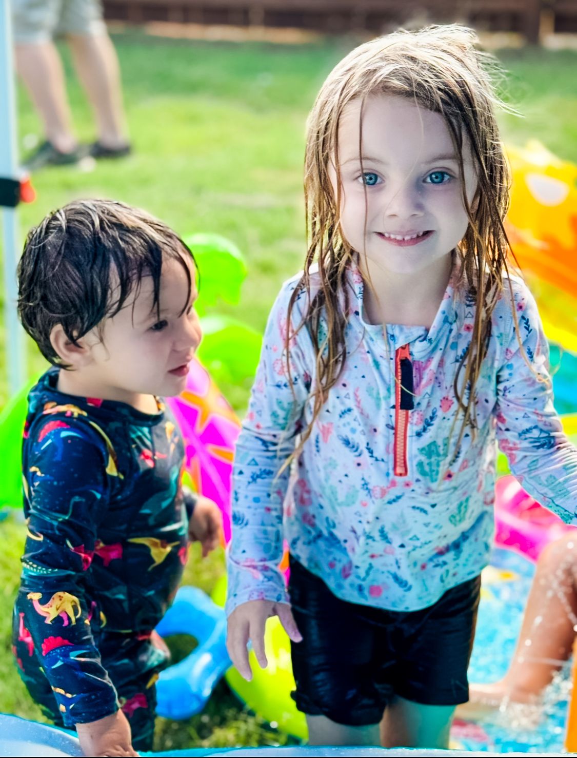 Two children playing outdoors with wet hair and colorful clothing on a grassy lawn.