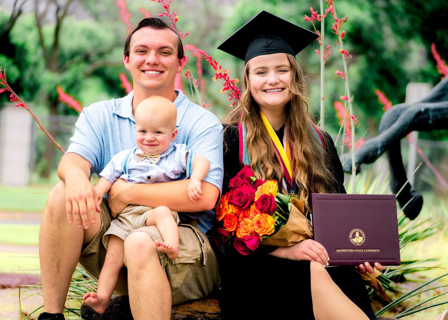 Graduation celebration photo outdoors with flowers and diploma in a garden setting.