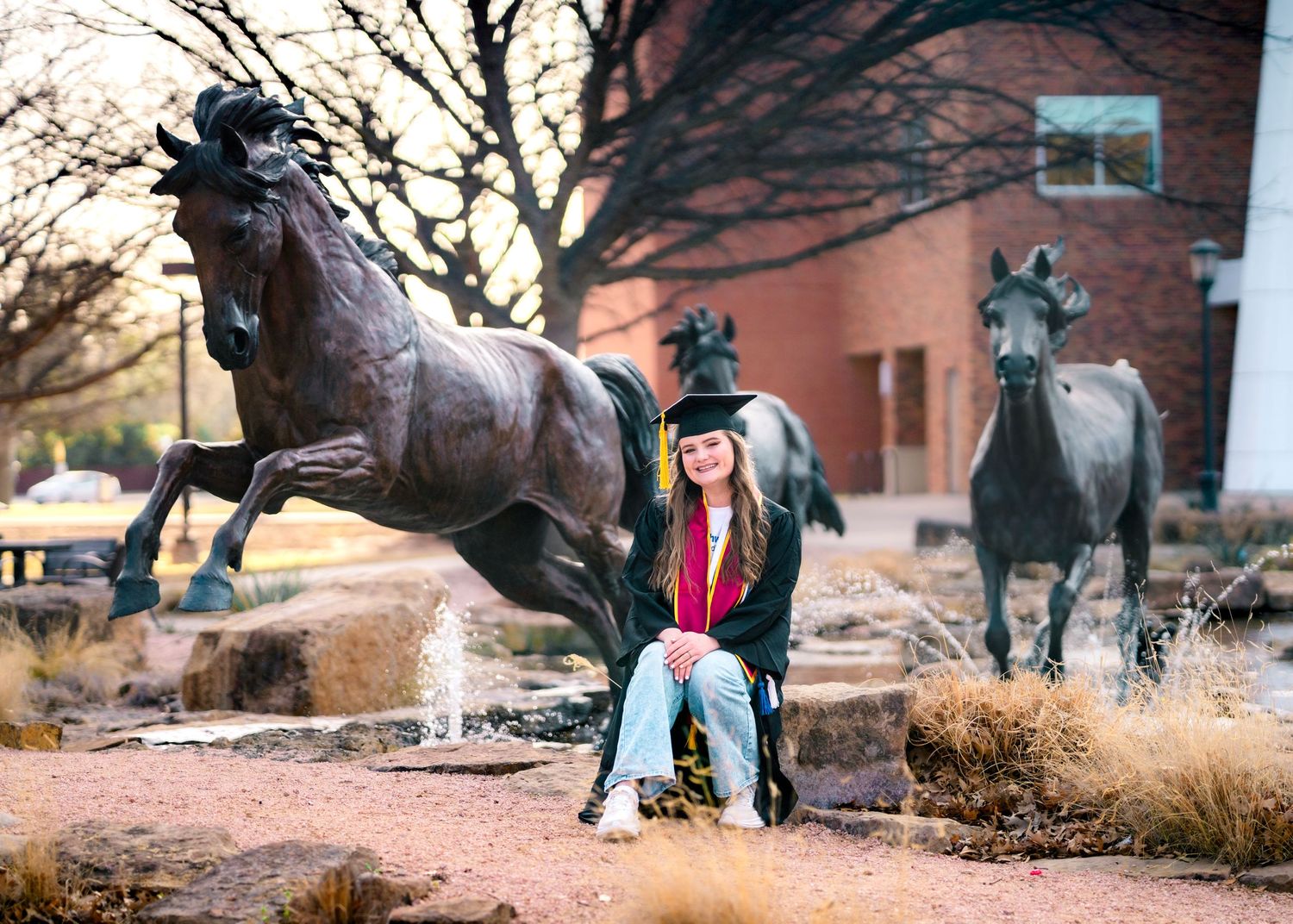 Graduate sitting on bronze horse statue outdoors wearing graduation cap and casual attire.