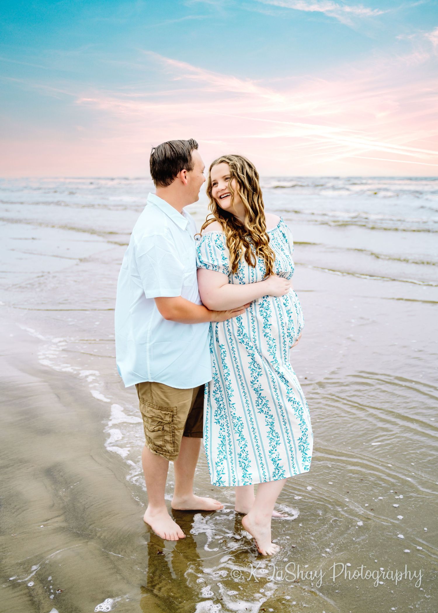A couple embraces during sunset on a beach while wearing coordinating blue and white outfits.