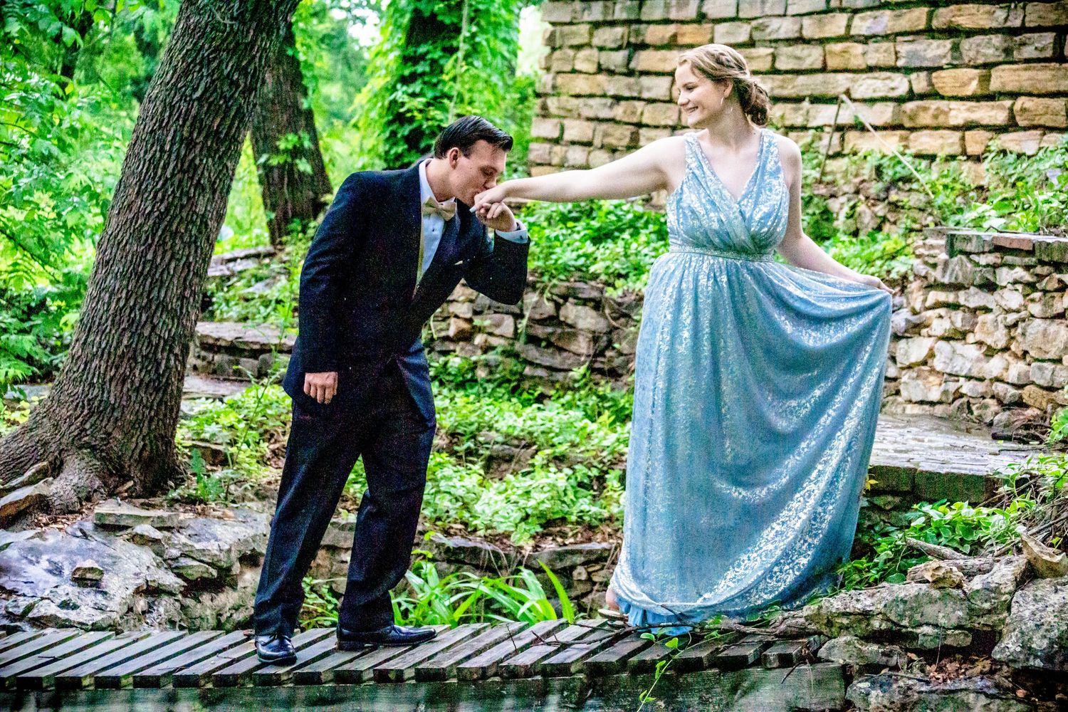 Couple in formal wear poses near stone wall in lush garden setting.