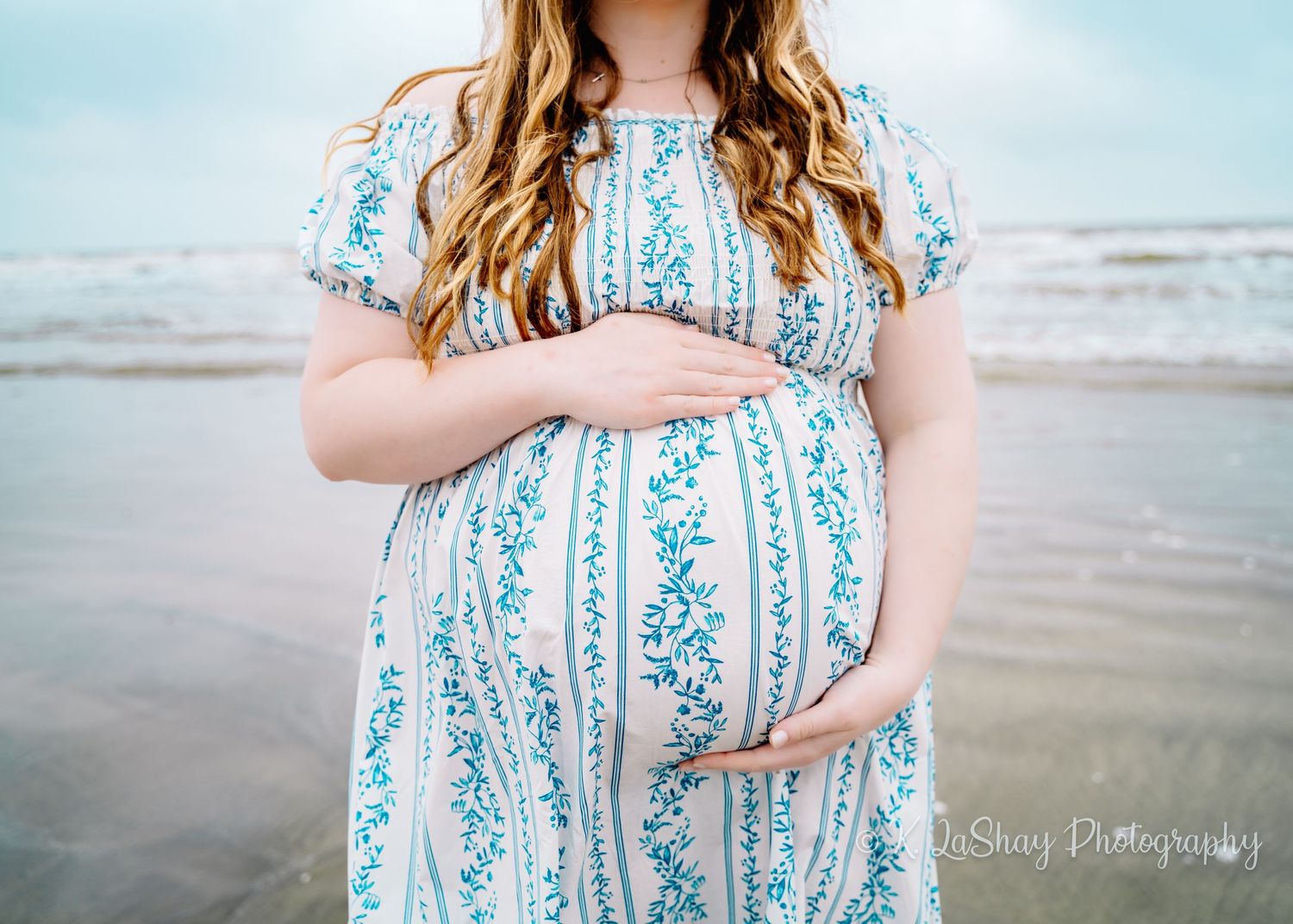 A maternity photo showing a blue and white striped dress against a beach backdrop.