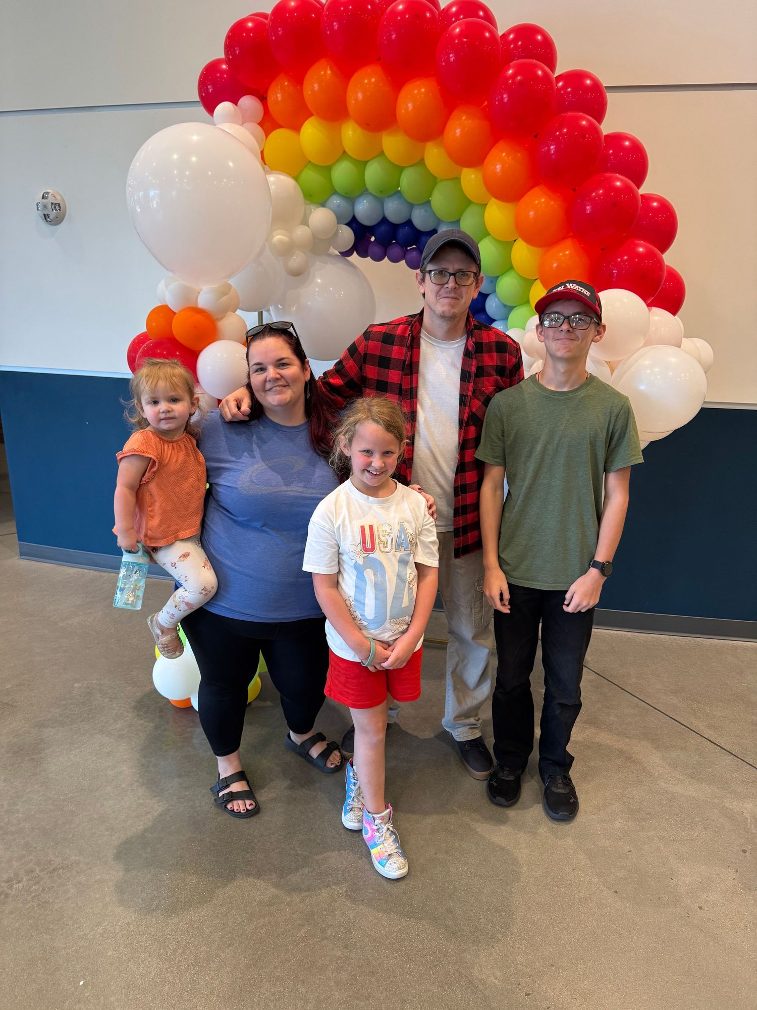 Group standing together under a vibrant rainbow balloon arch at an indoor event.