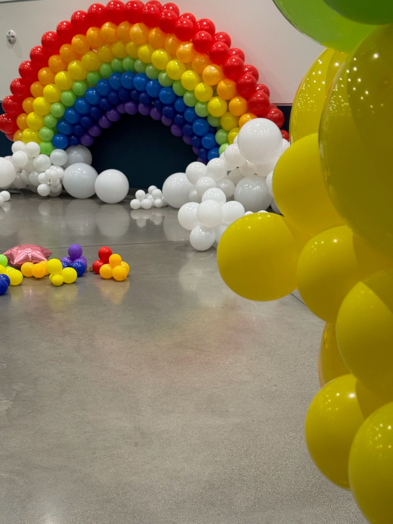 Rainbow colored balloon arch with yellow accents on concrete floor.