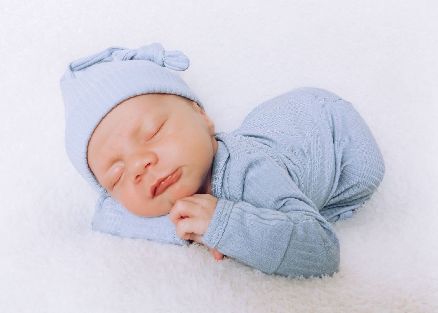 A sleeping newborn baby wearing a light blue outfit and matching cap on white bedding.