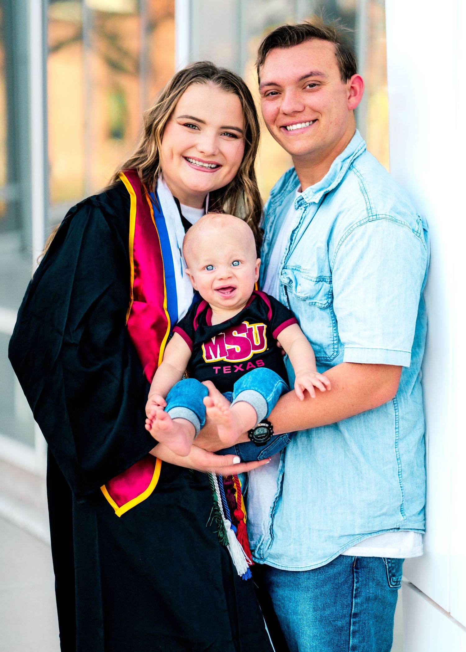 A couple in graduation attire poses together with a baby for celebratory photos outdoors.