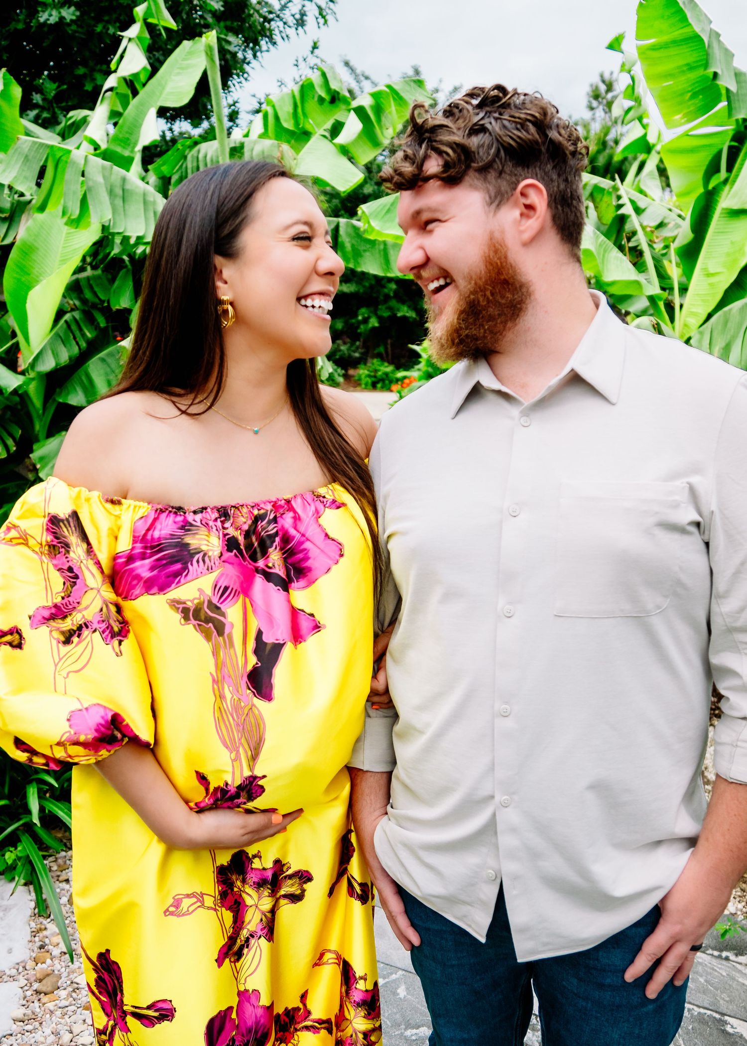 Couple poses together against tropical backdrop with yellow floral dress and white shirt.