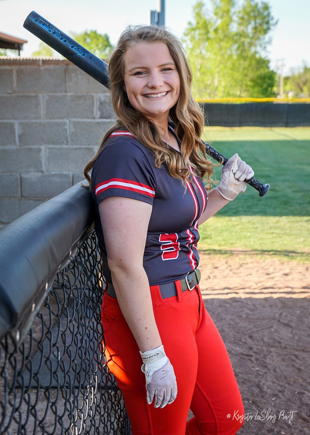 Softball player in red uniform pants and black shirt posing with bat against dugout fence.