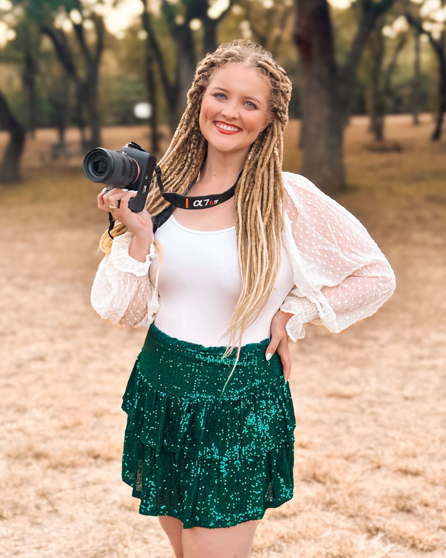 Photographer with blonde dreadlocks wearing a white blouse and green velvet skirt holding a camera outdoors.