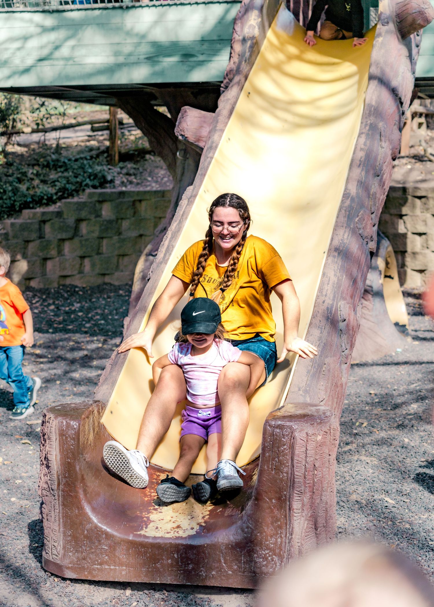 Adult and child enjoying a yellow playground slide together on a sunny day at a park.