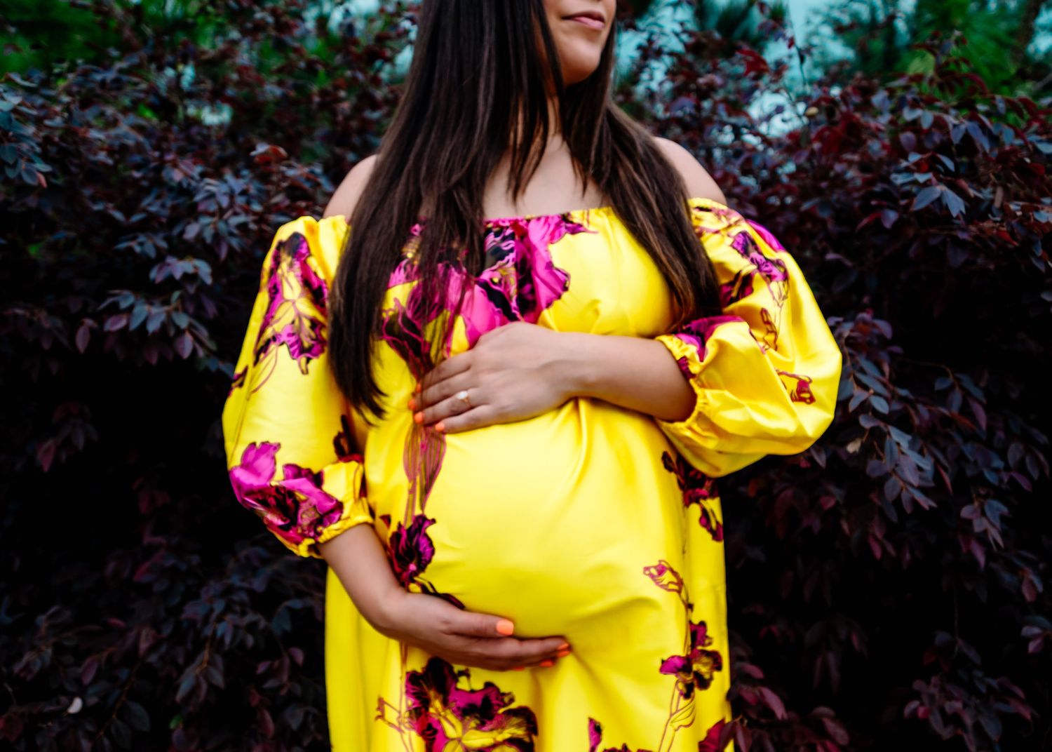 Yellow floral maternity dress contrasts beautifully against dark garden setting.