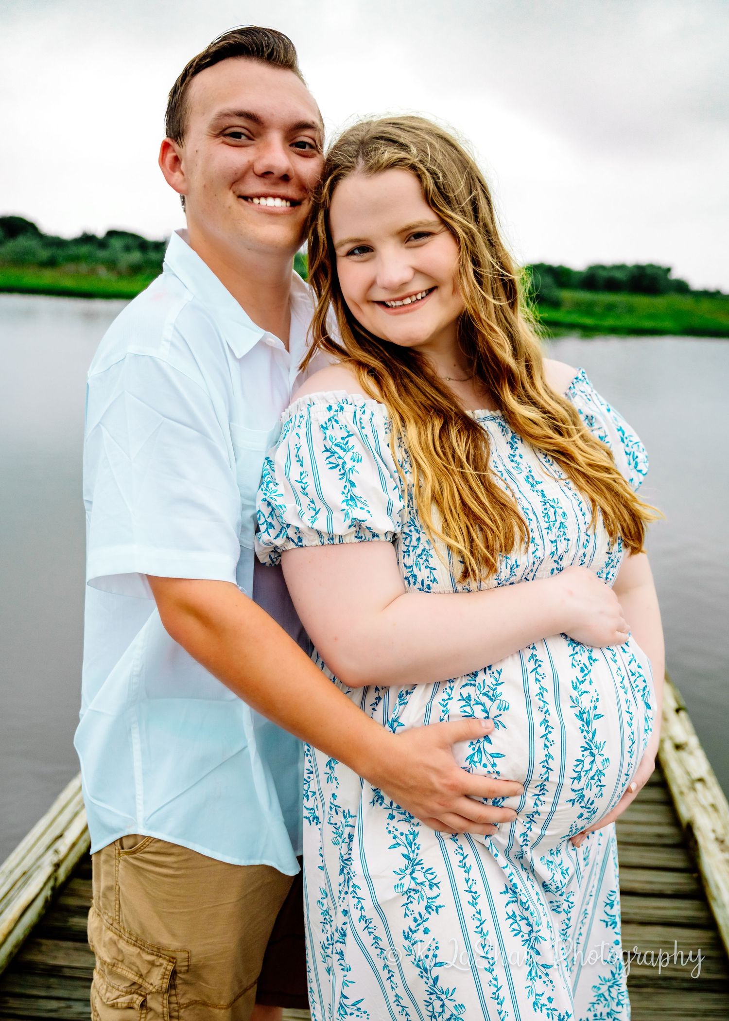 Happy couple embraces on a wooden pier with water and cloudy sky in background.