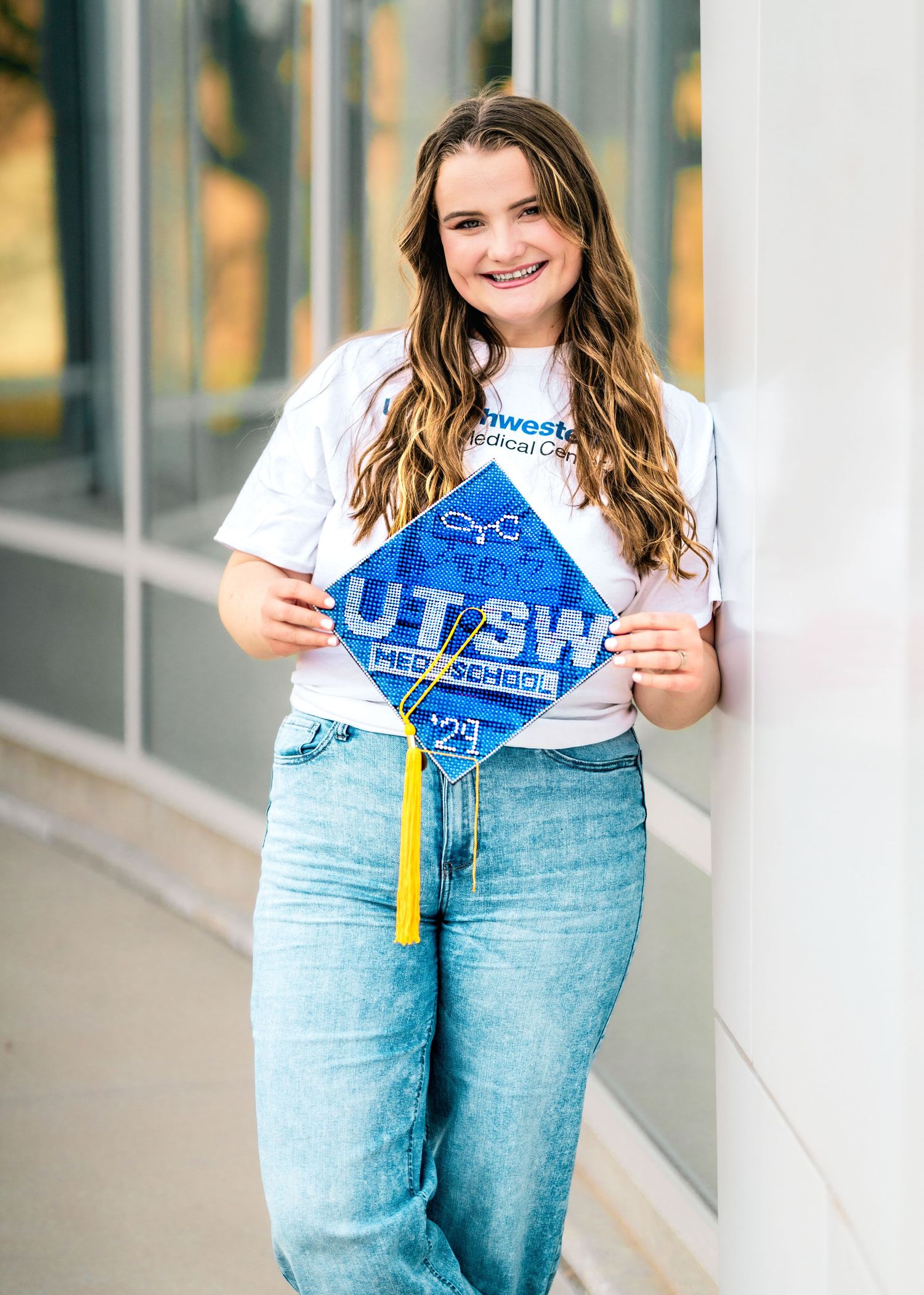 Graduate holding decorated blue cap while posing against light colored building exterior.