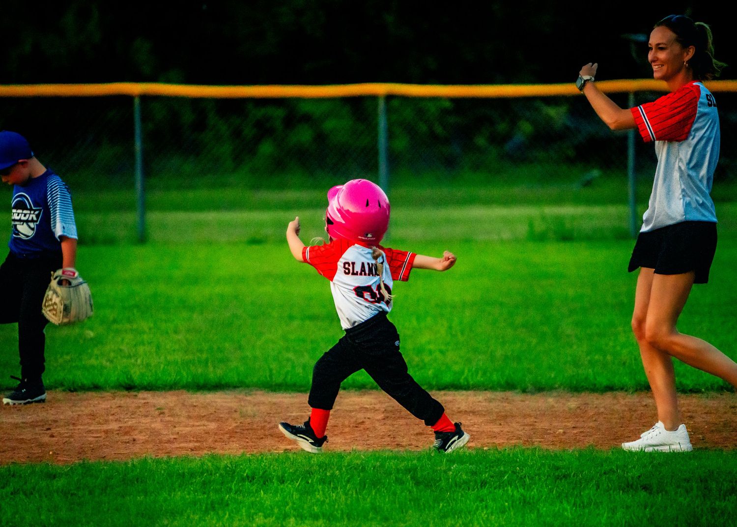 Children run and play on a baseball field with green grass and fencing.