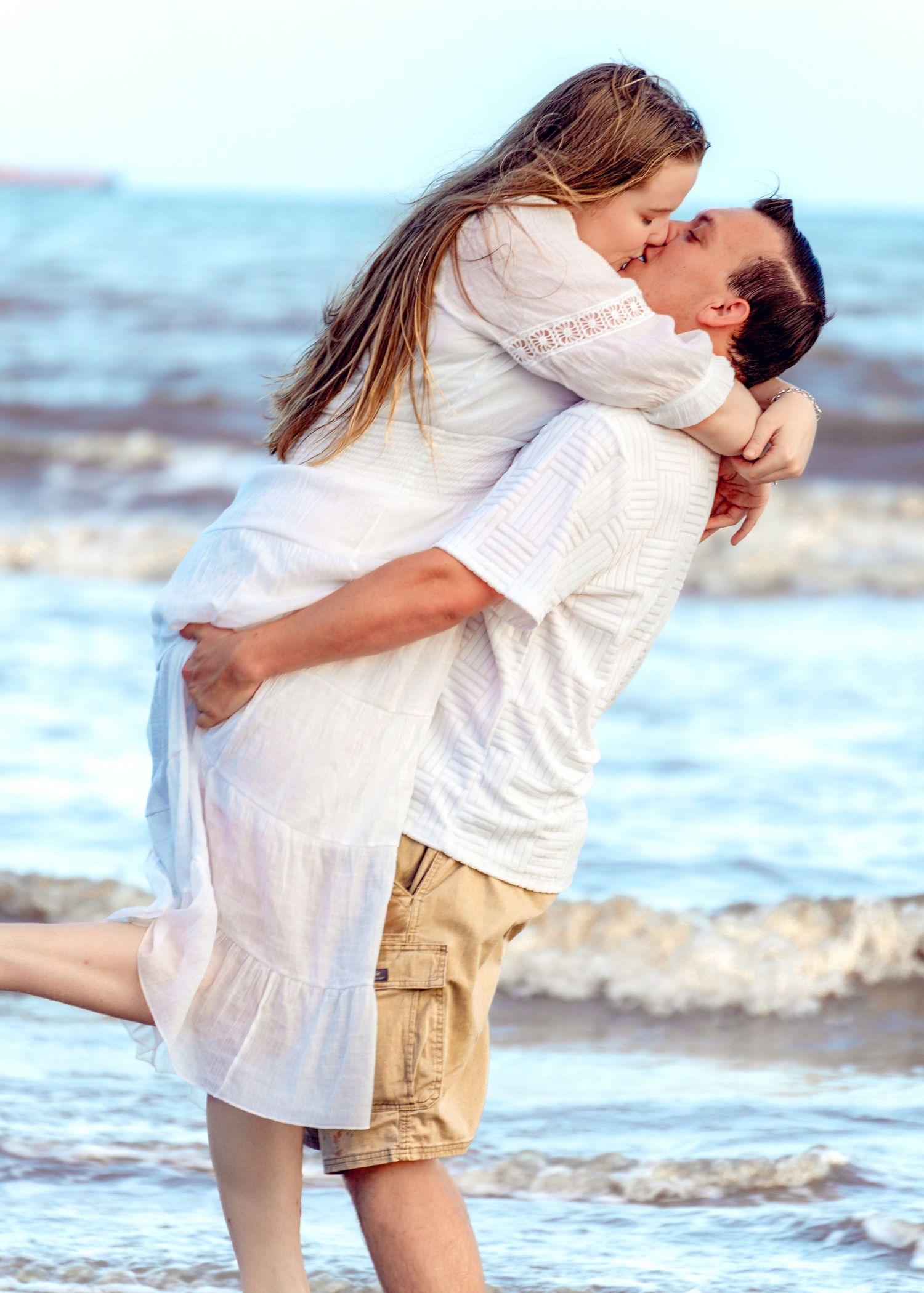 Couple embraces on beach with waves in background during sunset photoshoot.