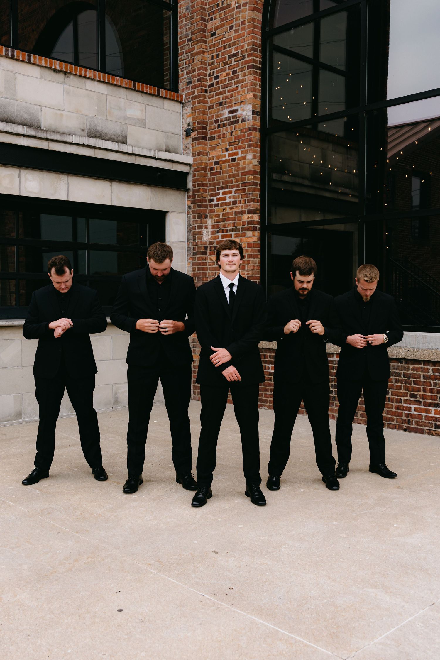 Groomsmen in black suits pose creatively lined up against building exterior.