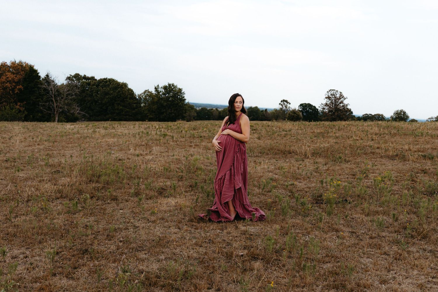 Figure in flowing burgundy dress walks through golden field at sunset.
