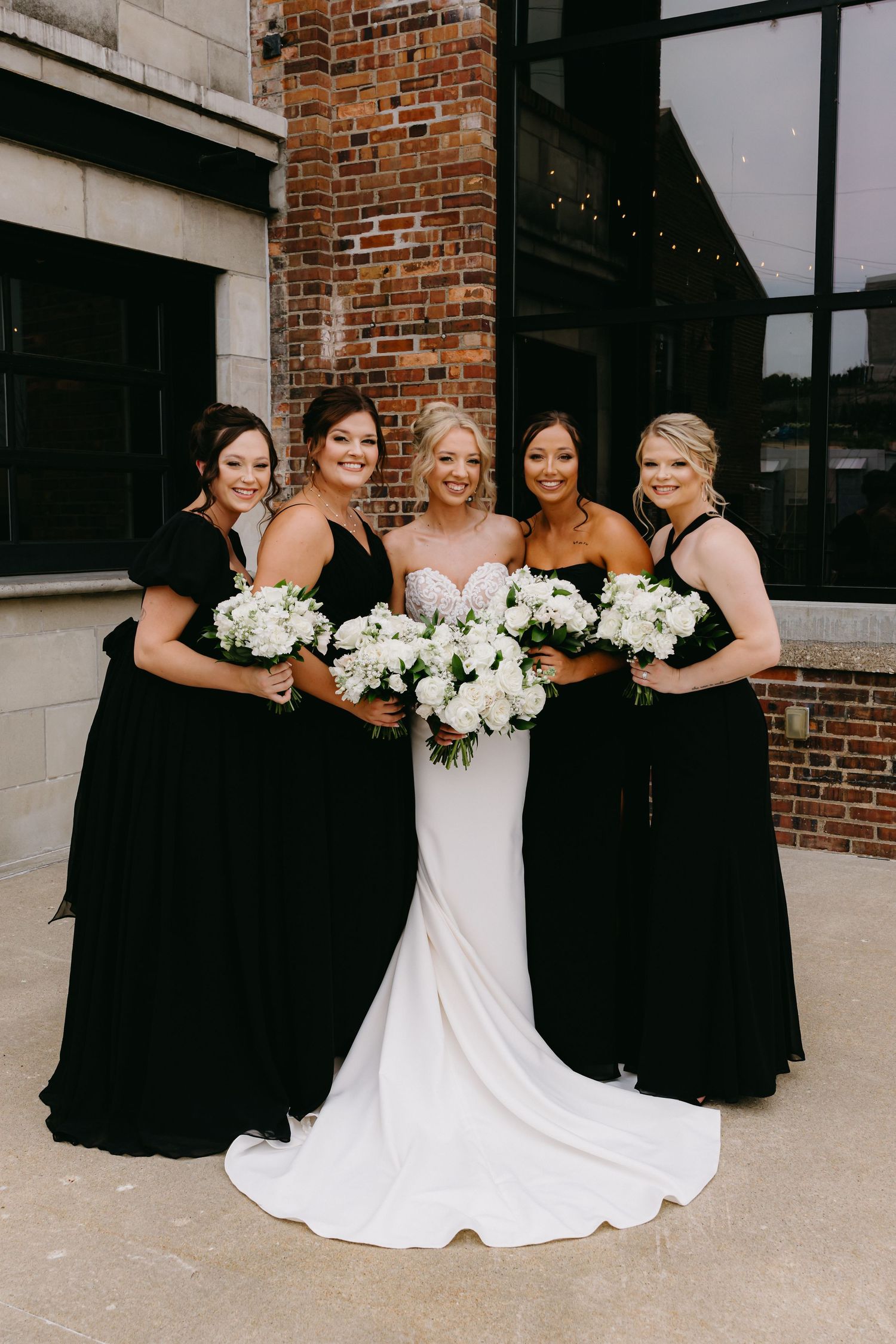Bridal party in black dresses and white gowns pose with bouquets against brick building.