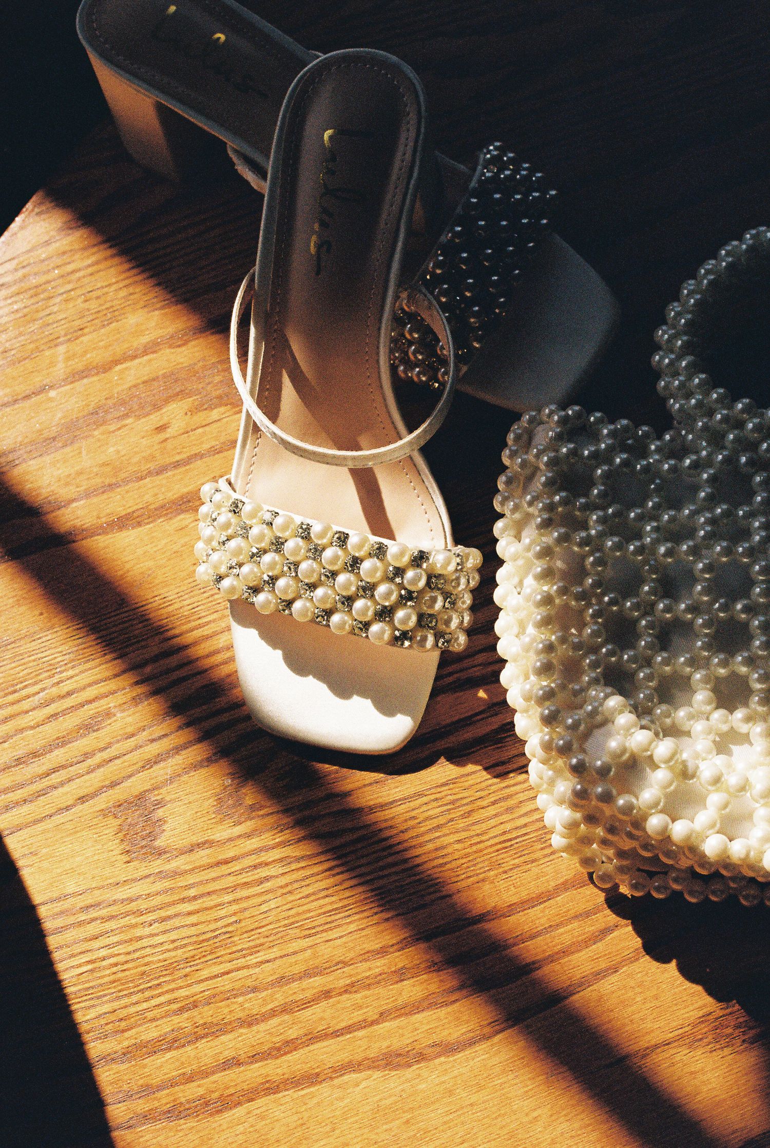 Pearl-embellished white sandal and matching handbag on wooden floor in dramatic lighting.