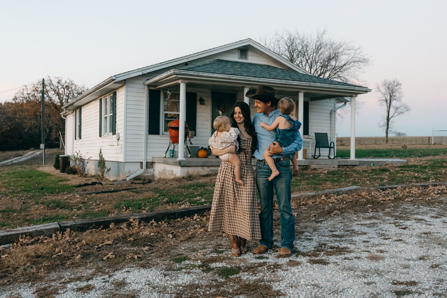 A family stands together outside a white farmhouse on a winter evening.