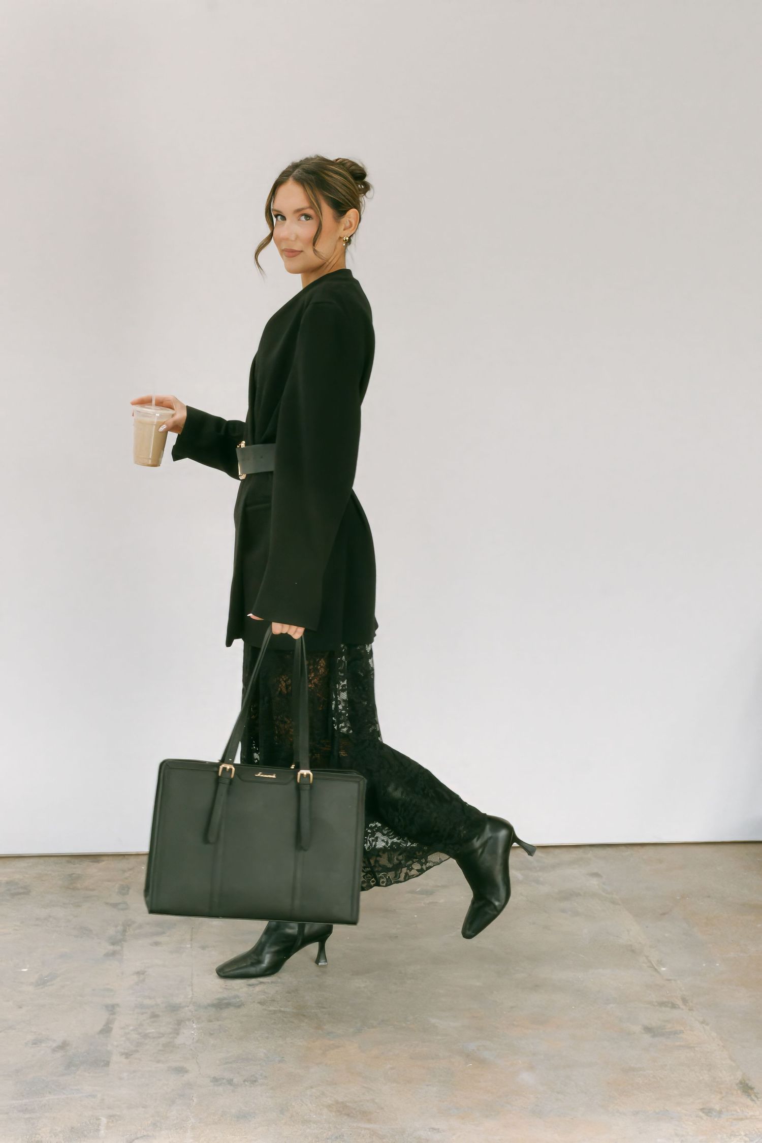 A person in all black attire walks across a minimalist space carrying a large dark leather tote bag.