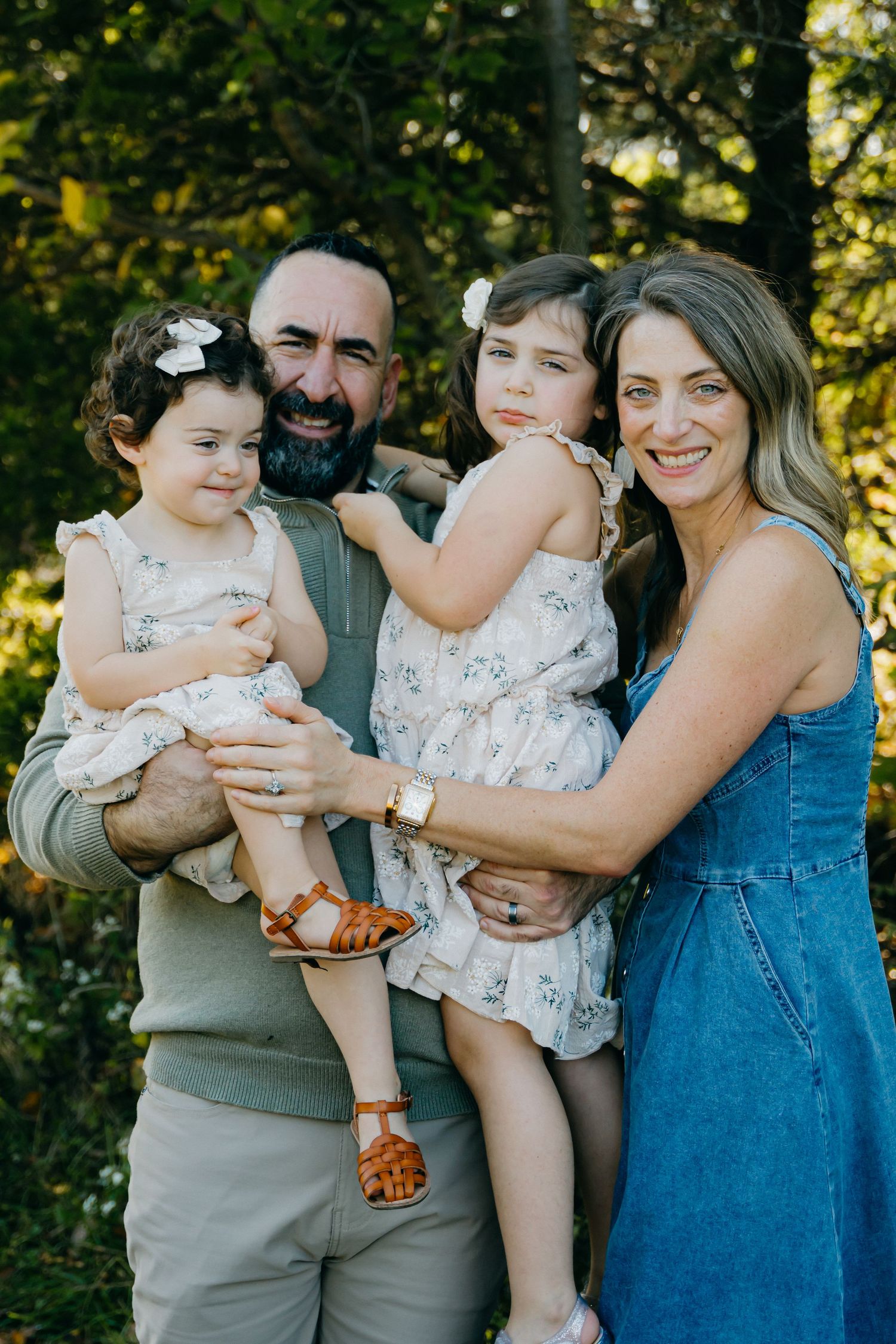 A joyful outdoor family portrait with matching floral dresses against a sunlit forest background.