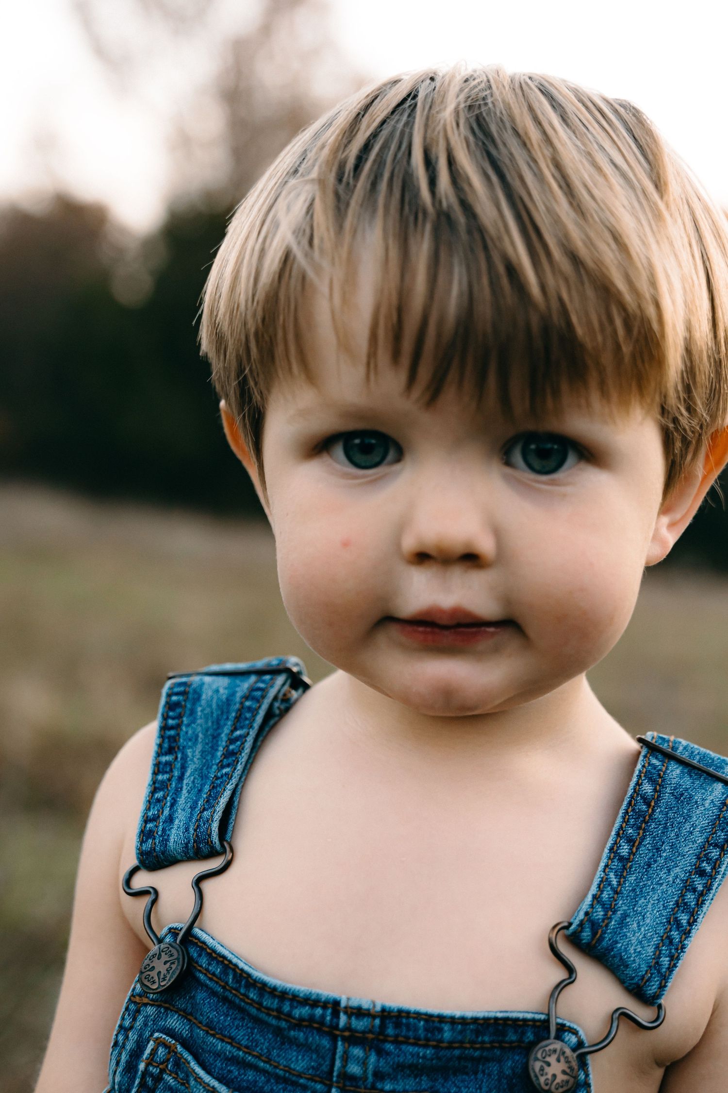 A toddler in denim overalls looks at the camera with bright blue eyes.