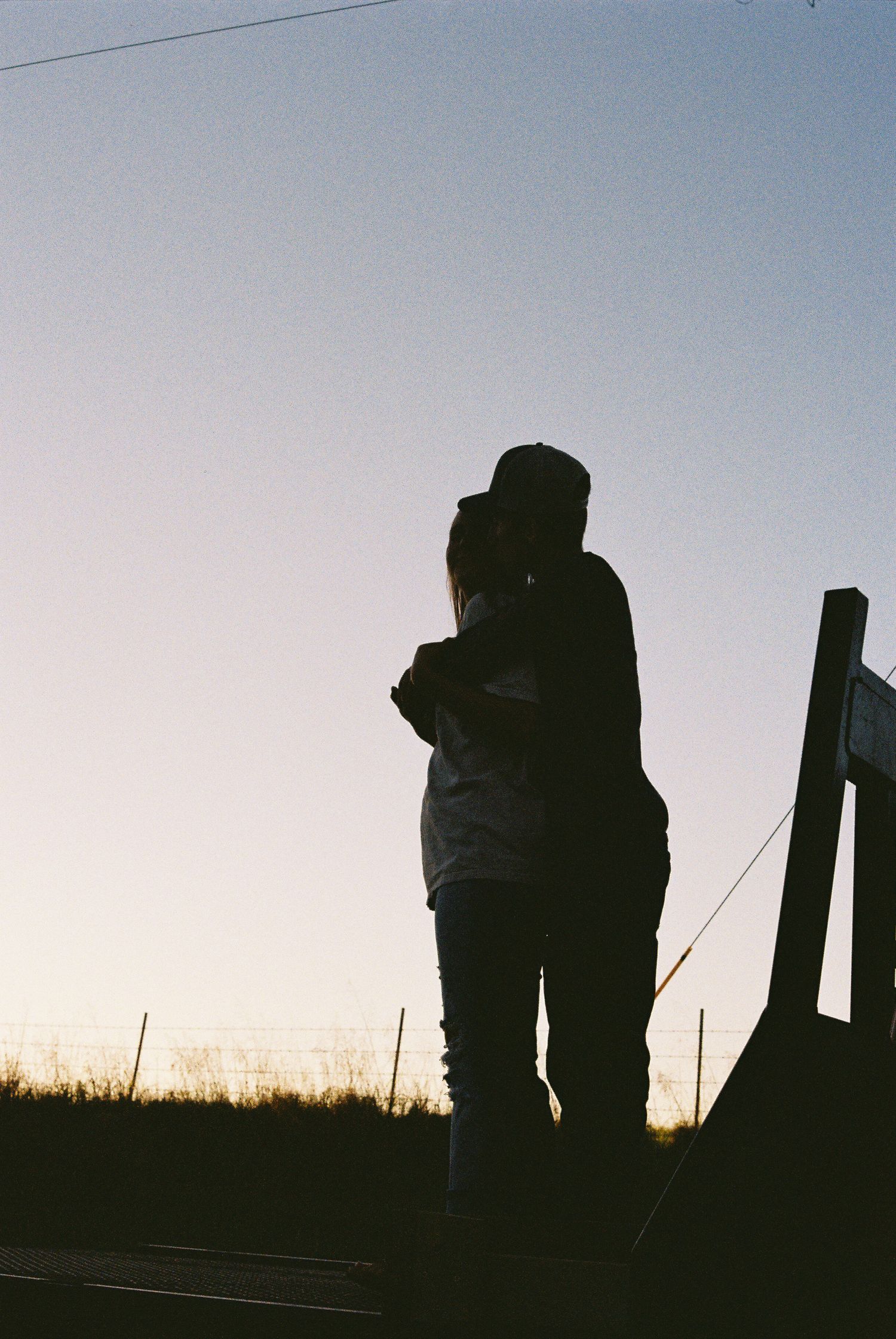Silhouetted figures embracing against a dusky sky with fence post.