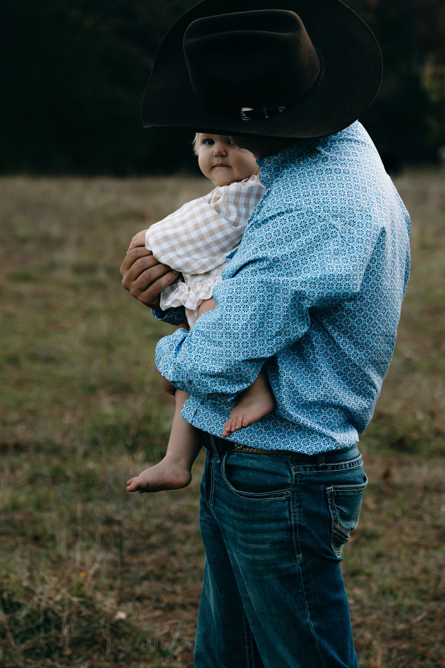 Someone in a cowboy hat holds a small child wearing gingham against a dusky field backdrop.