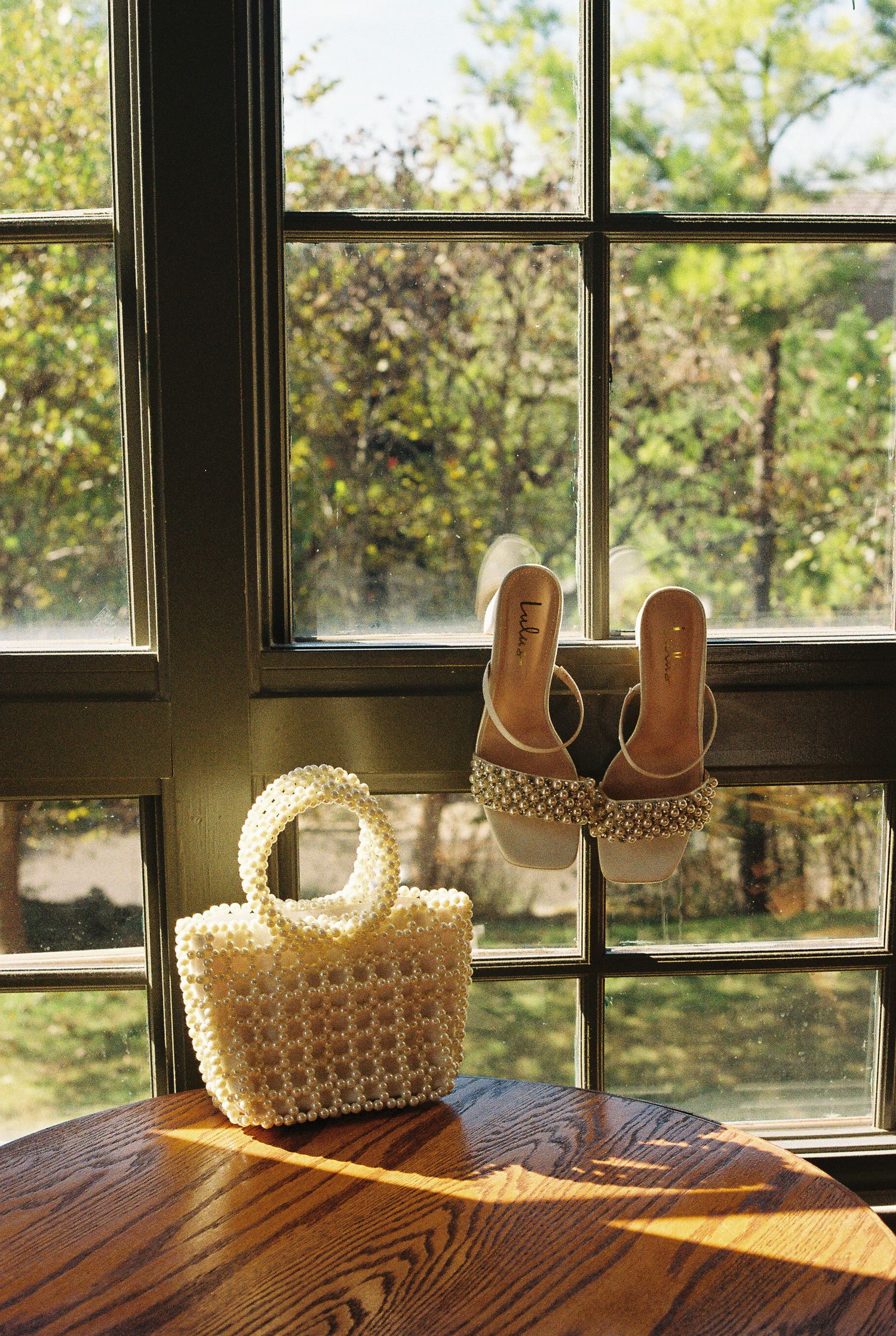 Pearl-beaded handbag and sandals displayed on wooden windowsill with natural lighting.