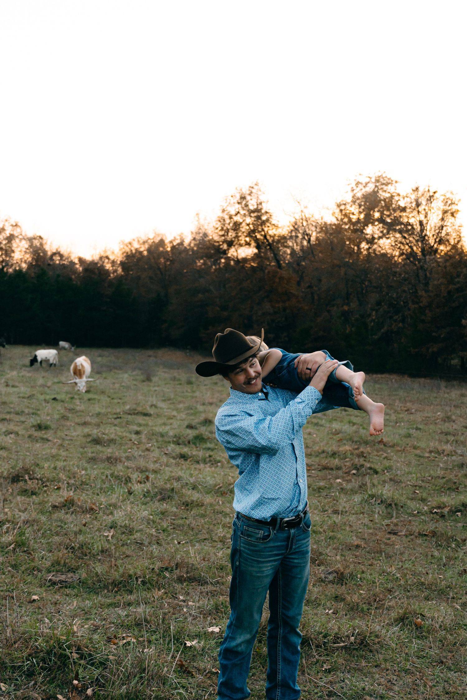 A person in western wear lifts a small child against a sunset pasture backdrop.