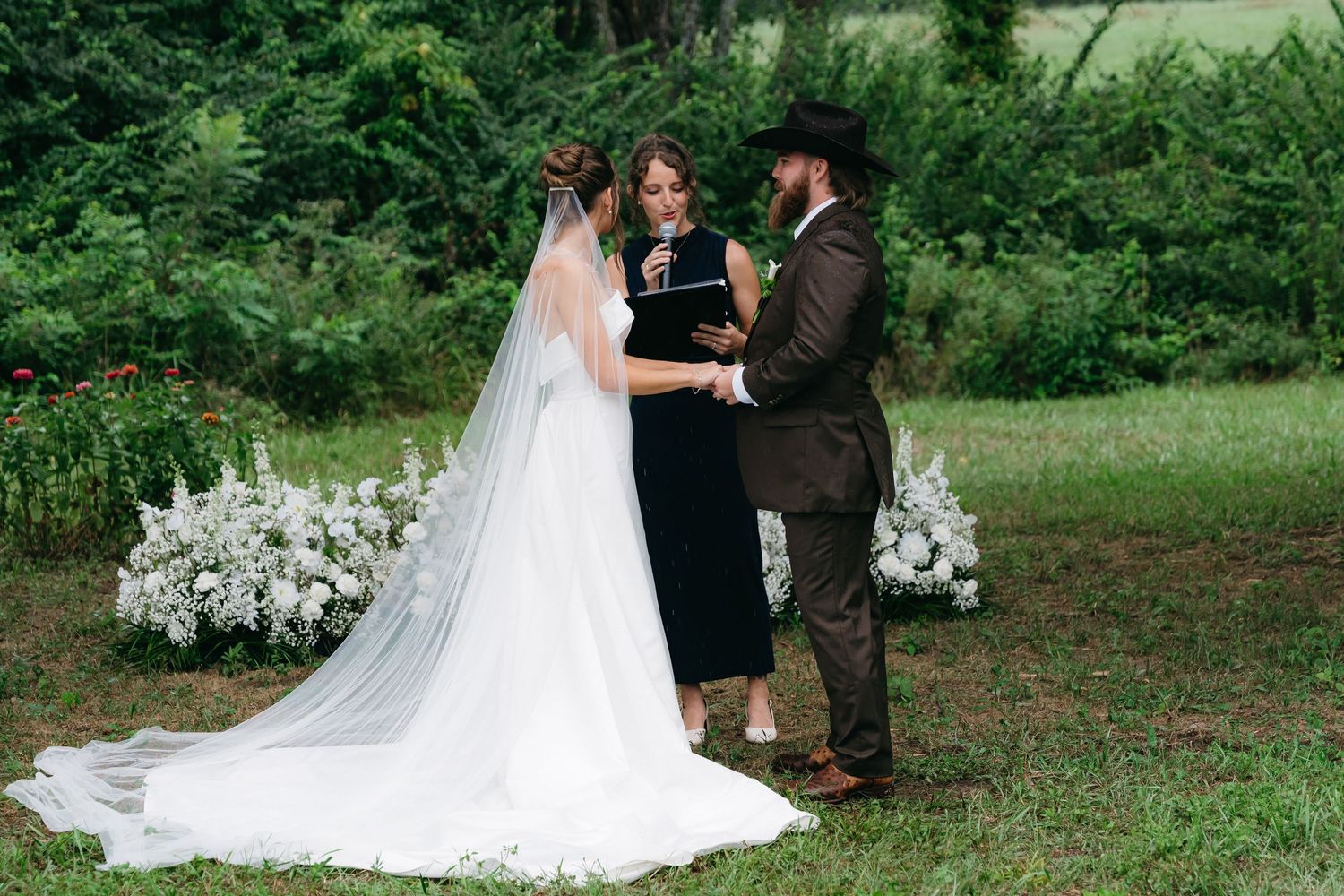 Outdoor wedding ceremony with flowing white dress against lush green background.