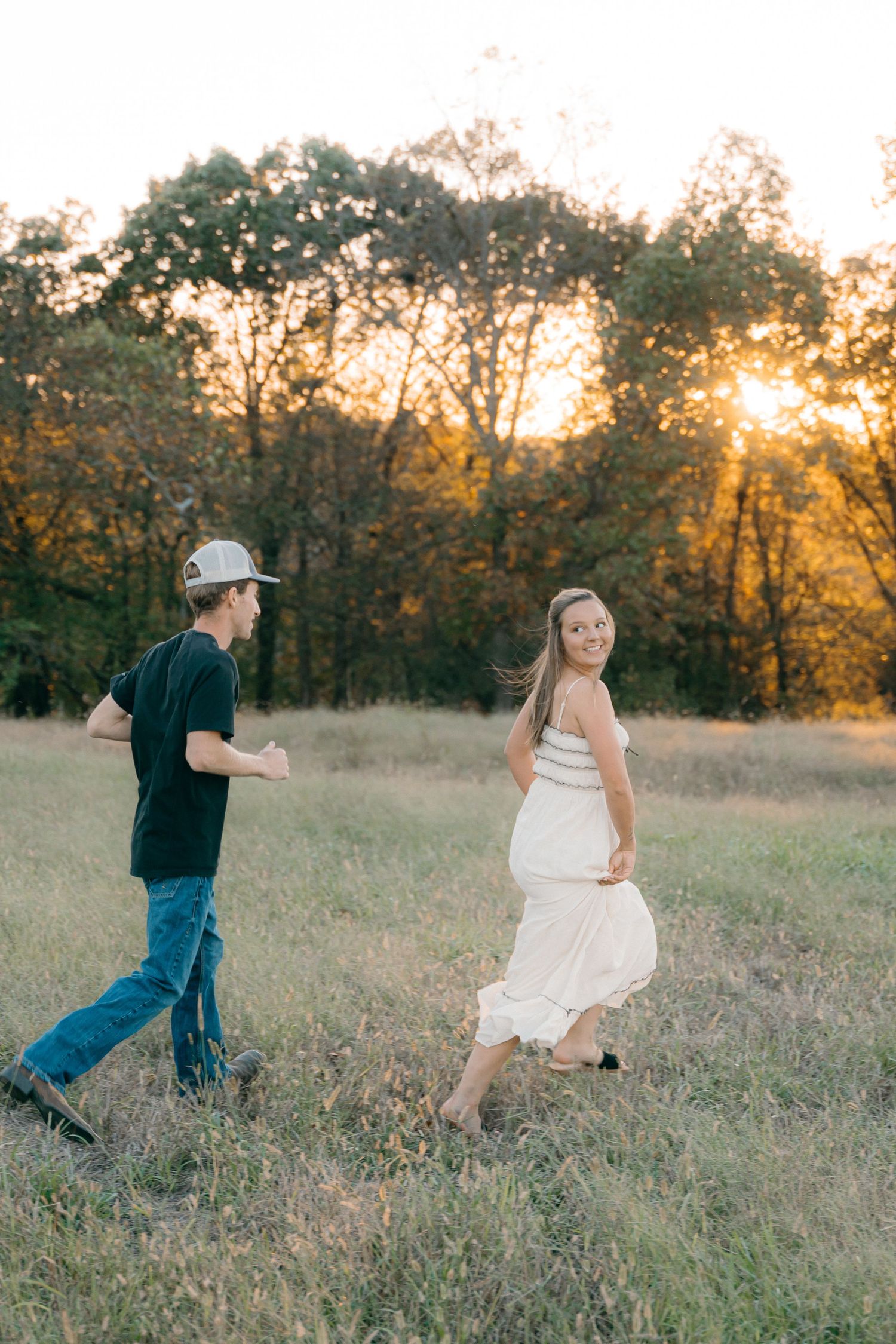 Playful couple running through a grassy field at sunset.