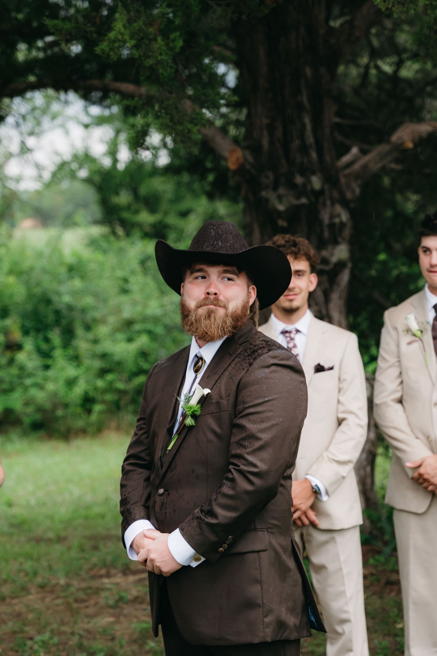 Wedding party in brown and beige suits standing outdoors under trees.