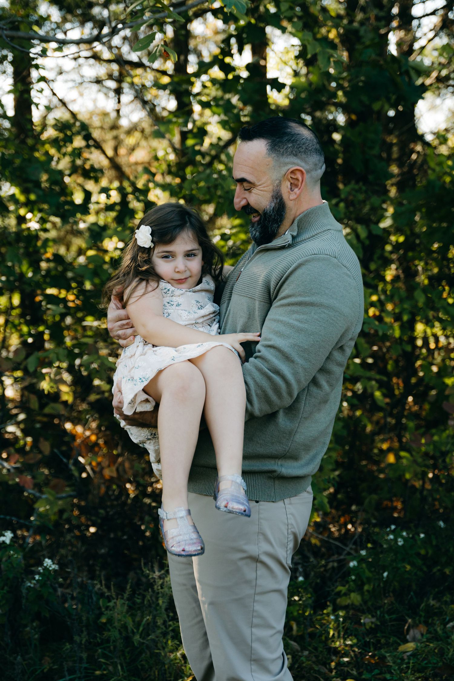 A heartwarming outdoor portrait showing a tender moment between parent and child against a sunlit forest backdrop.