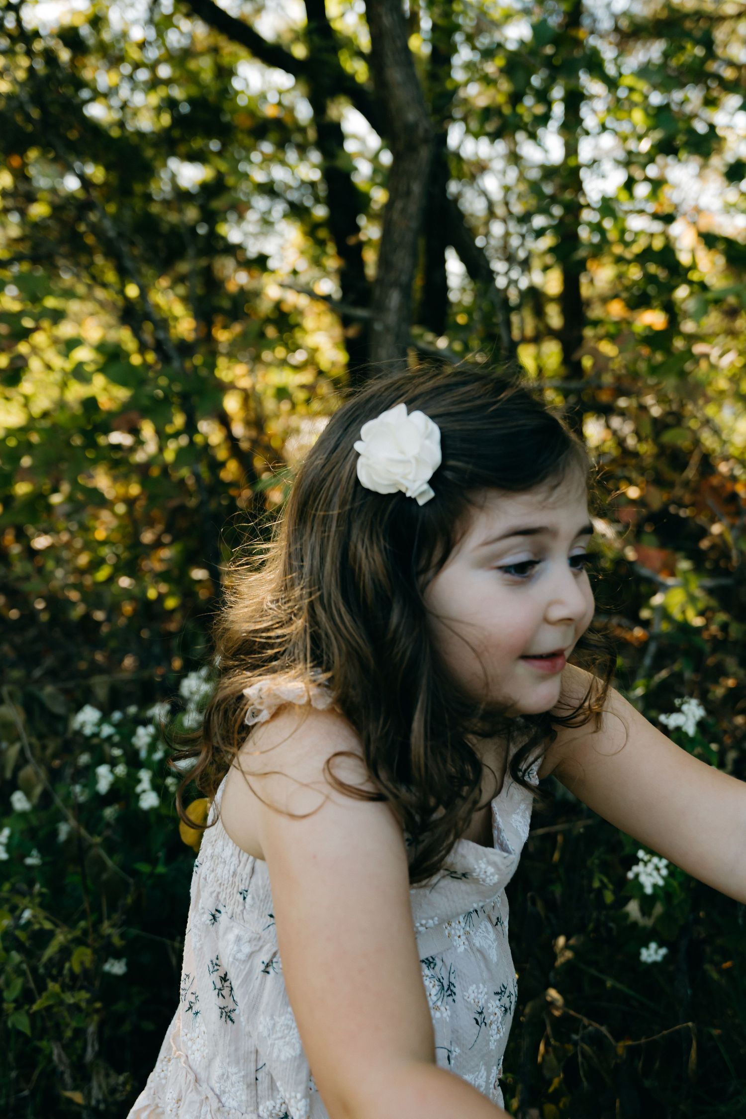 A child in a white floral dress plays in a wooded setting.