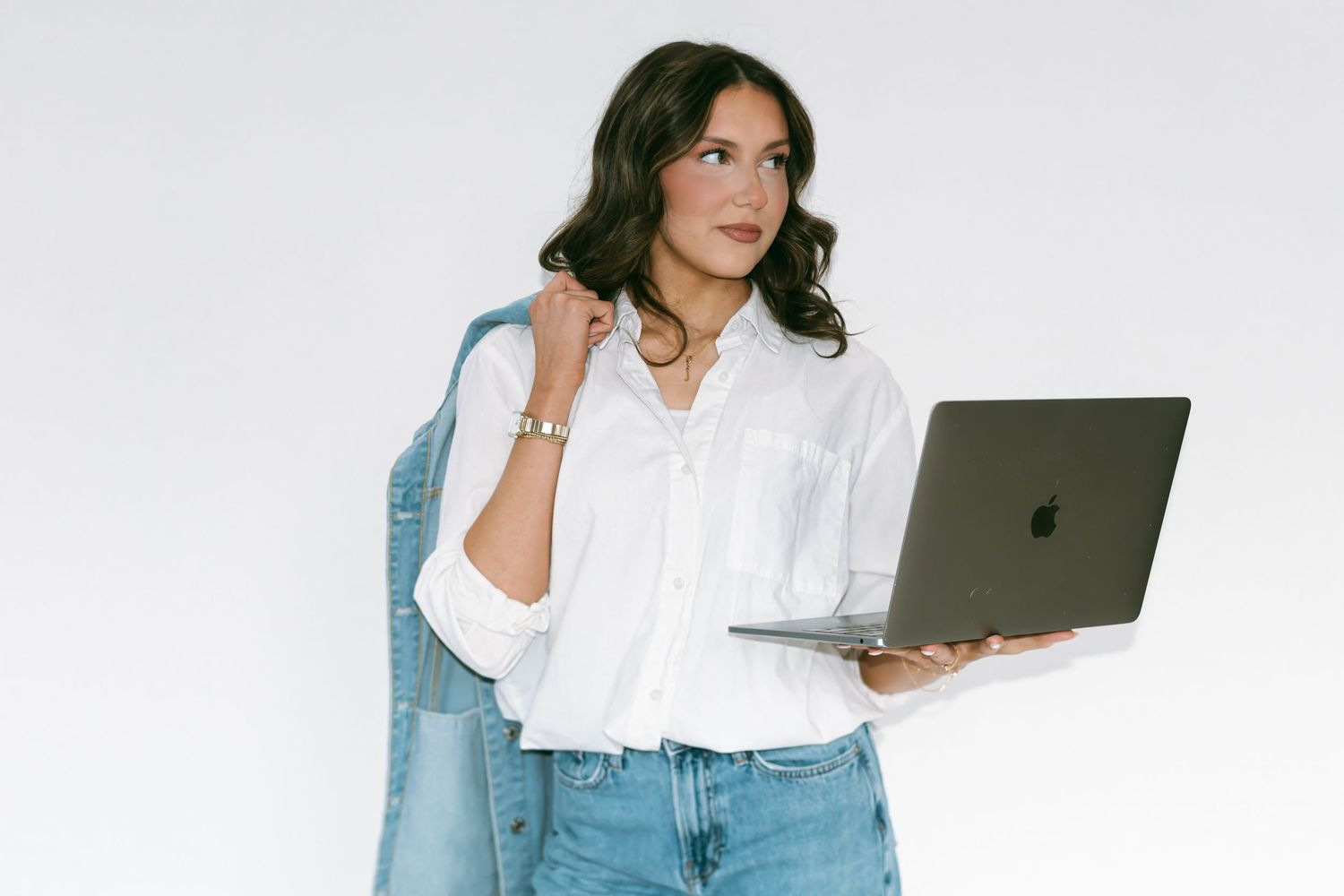 Professional in a white button-down shirt and jeans holds a MacBook laptop while standing against white background.